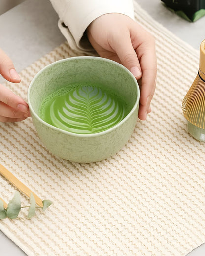 Green ceramic bowl with matcha tea and a hand holding it on a textured surface.